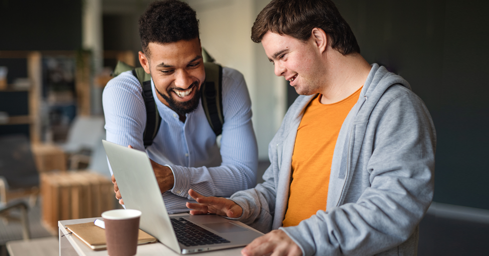 Auf dem Bild siehst du zwei junge Männer.
Sie gucken gemeinsam auf einen Laptop.
Die beiden Männer lachen.